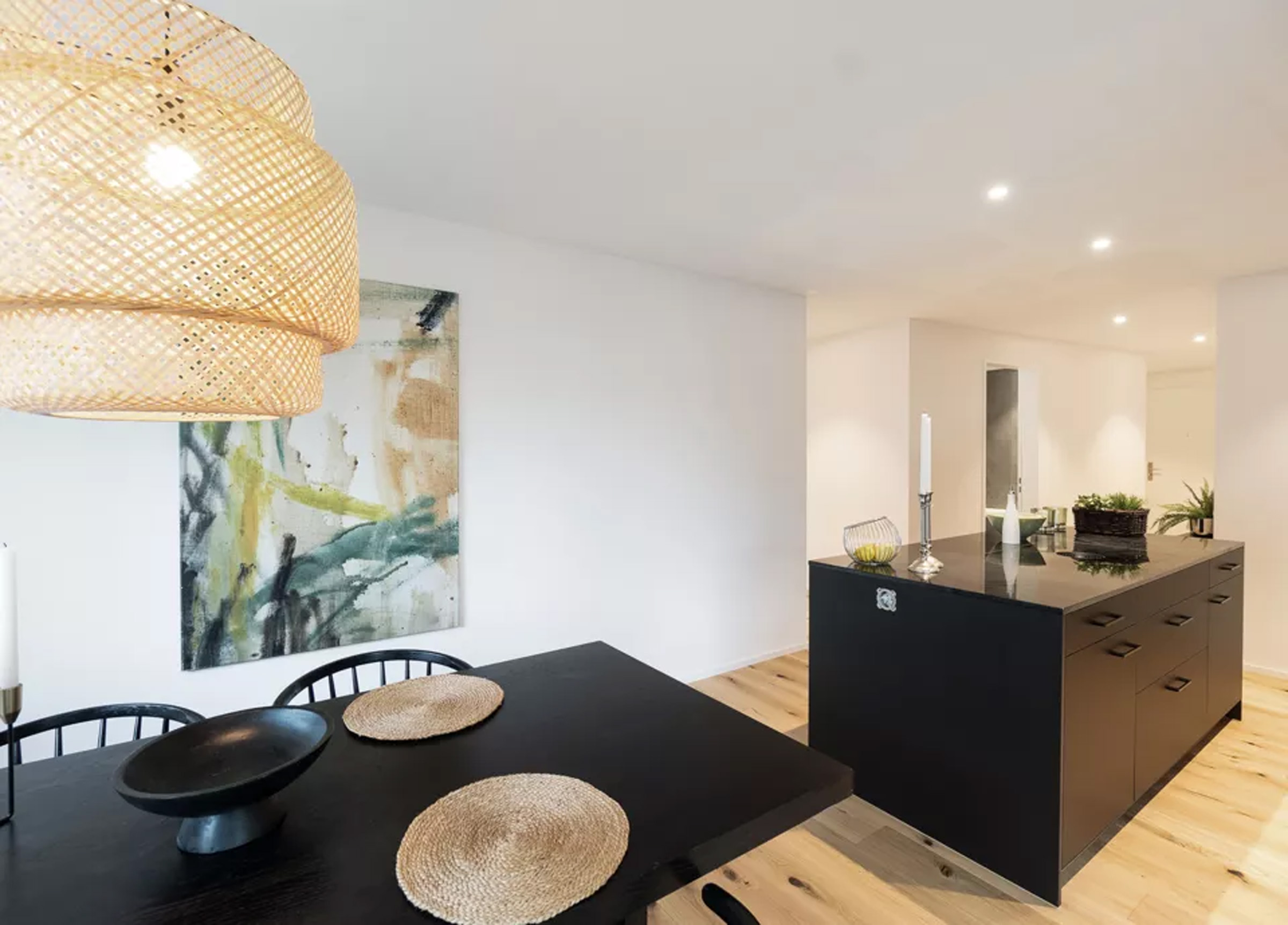Open-concept dining area with a black table, three woven placemats, and a large woven pendant light overhead; abstract art on a white wall behind.